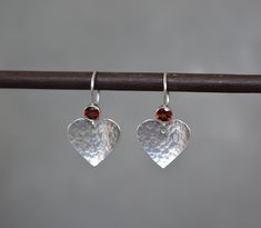 "Add a touch of love and sparkle to your jewelry collection with these delightful handmade sterling silver heart earrings. These elegant earrings feature  natural  faceted garnet gemstones and hand hammered hearts. The hammered dimpled surface of the hearts creates a play of light and shadow, catching the light from every angle and adding a subtle shimmer to your look. The earrings have a bright and shiny polished finish, adding a touch of radiance to their appearance. Choose from a selection of Silver Birthstone Earrings For Valentine's Day, Valentine's Day Silver Earrings With Birthstone, Valentine's Day Silver Birthstone Earrings, Silver Heart Earrings With Birthstone For Gift, Handmade Sterling Silver Heart Earrings For Wedding, Sterling Silver Heart Earrings With Ear Wire For Wedding, Handmade Teardrop Sterling Silver Heart Earrings, Handmade Teardrop Heart Earrings In Sterling Silver, Handmade Sterling Silver Teardrop Heart Earrings