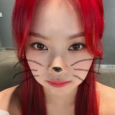 a girl with red hair and cat makeup looks at the camera while she has her face painted to look like a cat