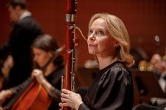 a woman in black shirt holding a musical instrument and looking off into the distance with orchestra behind her