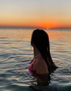 a woman in the water at sunset with her back to the camera, looking out into the distance