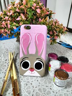 an iphone case with a bunny face painted on it next to paintbrushes and flowers