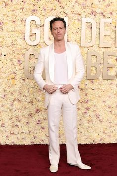 a man in a white suit and tie standing on a red carpet with flowers behind him