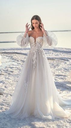 a woman standing on top of a sandy beach wearing a wedding dress with long sleeves