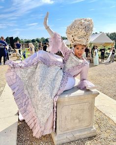 a woman sitting on top of a stone block in a dress and hat with her arms outstretched