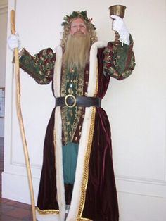 a bearded man dressed in costume holding a cup and standing next to a white wall