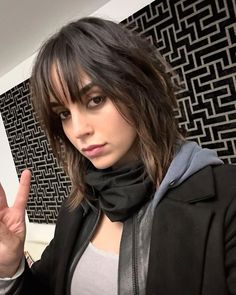 a woman with long hair wearing a black jacket and gray shirt is making the peace sign