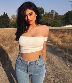 a woman wearing jeans and a crop top is standing in the middle of a dirt road