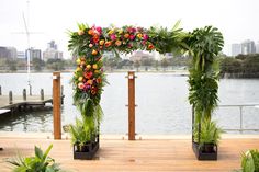 an arch made out of plants and flowers on a wooden platform near the water with buildings in the background