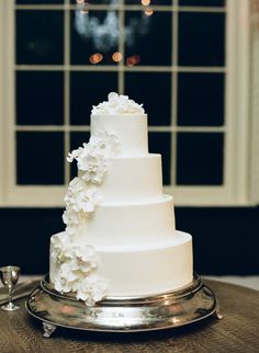 a wedding cake with white flowers on top sits on a table in front of a window