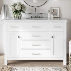 a white bathroom vanity with a round mirror above it and flowers on the counter top