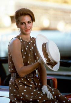 a woman sitting on the back of a car holding a white polka dot hat in her right hand