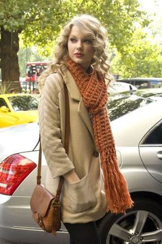 a woman standing next to a parked car wearing a scarf and purse with her hands in her pockets