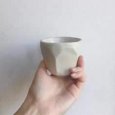 a hand holding a white ceramic cup in front of a white wall with no one around it