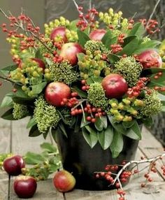 an arrangement of apples and berries in a pot on a wooden table with green leaves