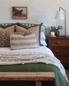 a bed with pillows and blankets on top of it next to a wooden bench in front of a painting