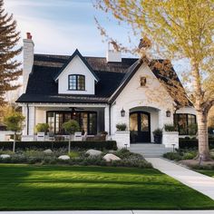 a white house with black shingles and trees in the front yard, surrounded by green grass