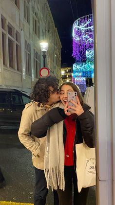 a man and woman kissing in front of a building with christmas lights on the street behind them