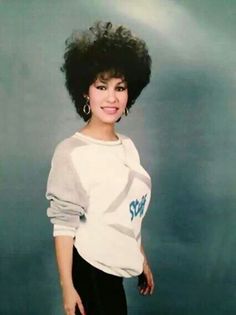 a woman with an afro standing in front of a blue background wearing a white shirt and black pants