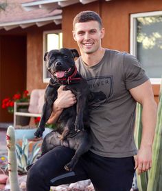 a man holding a black dog in his arms and smiling at the camera while standing next to a house