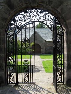 an ornate iron gate opens into a courtyard with green grass and stone buildings in the background