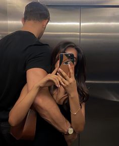 a man and woman standing next to each other in front of an elevator while looking at their cell phones