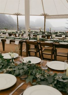 tables set up with white plates and greenery under a large tented area for an outdoor wedding