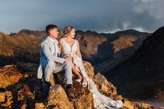 a bride and groom sitting on top of a mountain looking at each other with mountains in the background