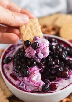 a hand holding a cracker over a bowl of blueberry cheesecake dip