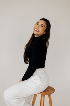 a woman sitting on top of a wooden stool wearing white pants and a black shirt