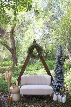 a white couch sitting in the middle of a forest next to a wooden structure with a wreath on it