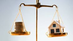 a scale with two houses on top of each other and coins stacked in front of them