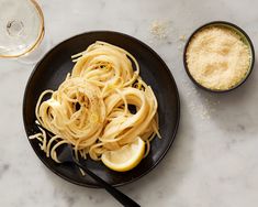 a black plate topped with pasta and lemon wedges next to a bowl of grated parmesan cheese