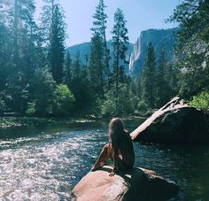 a woman sitting on top of a rock in the middle of a river surrounded by trees