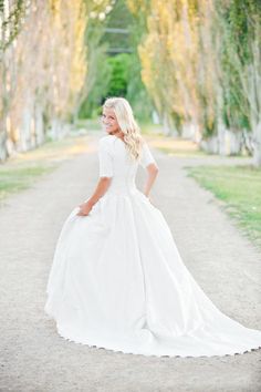 a woman in a white wedding dress standing on a dirt road with trees behind her