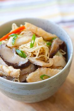 a white bowl filled with noodles and meat on top of a wooden table next to carrots