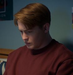 a young man in a red shirt looking at his cell phone while sitting on a bed