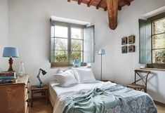 a bed sitting under two windows next to a wooden table and chair in a bedroom