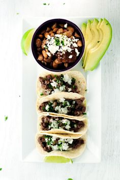 three tacos on a plate with beans, avocado and sour cream in the background