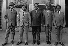 five men in suits and ties standing next to each other on the sidewalk near a building