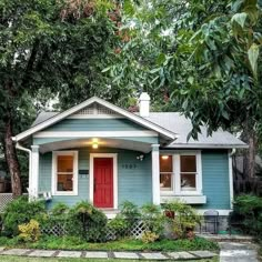 a small blue house with red door and windows in the front yard is surrounded by greenery