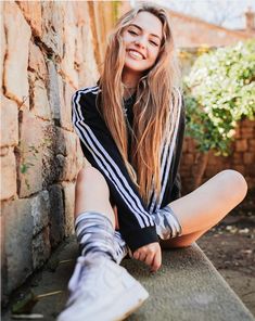 a young woman sitting on the ground next to a brick wall wearing adidas shoes