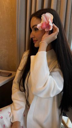 a woman with long black hair holding a flower in her hair and wearing a white sweater