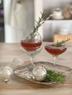 two glasses filled with liquid and garnished with rosemary on a silver tray next to christmas ornaments