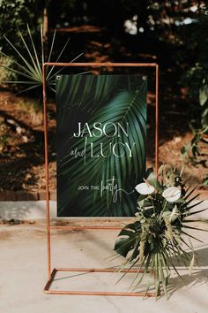 a sign that is sitting on the ground with flowers in front of it and greenery behind it