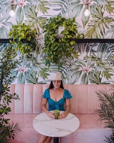 a woman sitting at a table in front of a wall with plants and potted plants