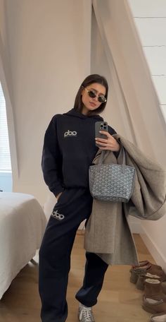 a woman standing in front of a bed holding a purse and looking at her cell phone