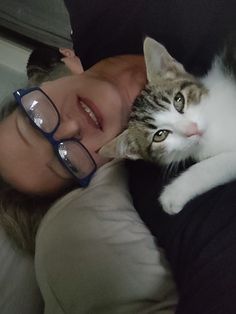 a woman laying on top of a bed with a cat next to her face and wearing glasses