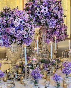 purple flowers are arranged in silver vases on a table with candles and other decorations