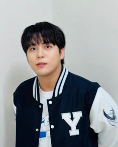 a young man wearing a varsity jacket and white t - shirt, standing in front of a wall