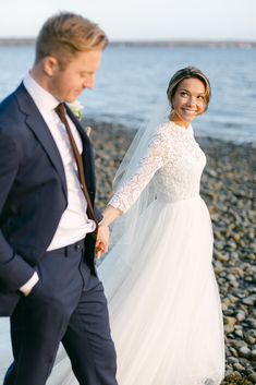 a bride and groom are walking on the beach holding hands while smiling at each other